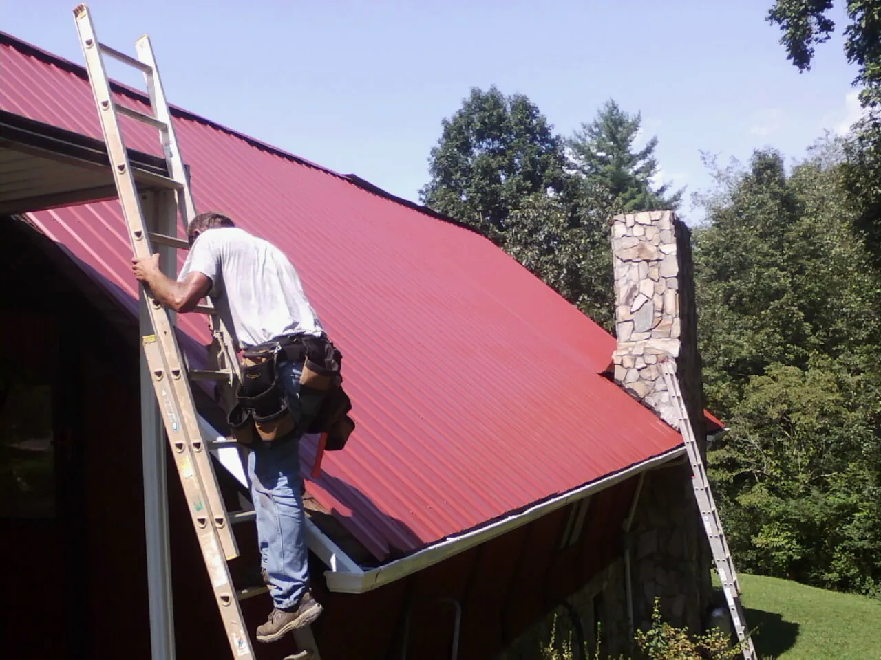 Roofer on a ladder beside a red metal roof and stone chimney — Soffit & Fascia Repair by Roofing Brothers in Ennis